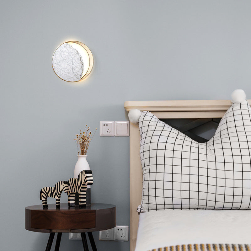 Marble bedside LED wall lamp in circular design, mounted above a wooden side table with decorative items next to a bed with a checkered pillow.